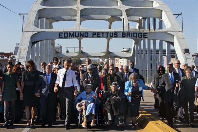 President Barack Obama, fourth from left, walks holding hands with Amelia Boynton Robinson, who was beaten during \”Bloody Sunday,\” as they and the first family and others including Rep. John Lewis, D-Ga, left of Obama, walk across the Edmund Pettus Bridge in Selma, Ala,. for the 50th anniversary of the landmark event of the civil rights movement, Saturday, March 7, 2015. At far left is Sasha Obama and at far right is former first lady Laura Bush. Adelaide Sanford also sits in a wheelchair.