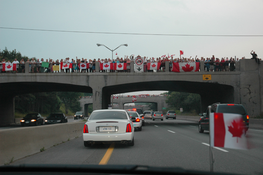 People wave flags along the Highway of Heroes as Jordan Anderson’s casket drives by.