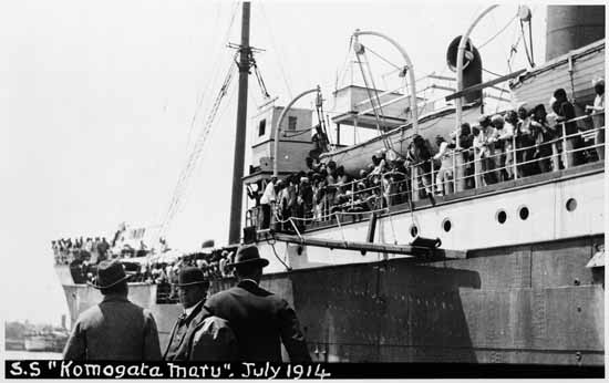 Three men are pictured in front of the Komagata Maru, with passengers on deck. July 1914.