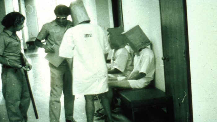 Two guards speak to several prisoners with bags on their heads during the 1971 Stanford Prison Experiment.