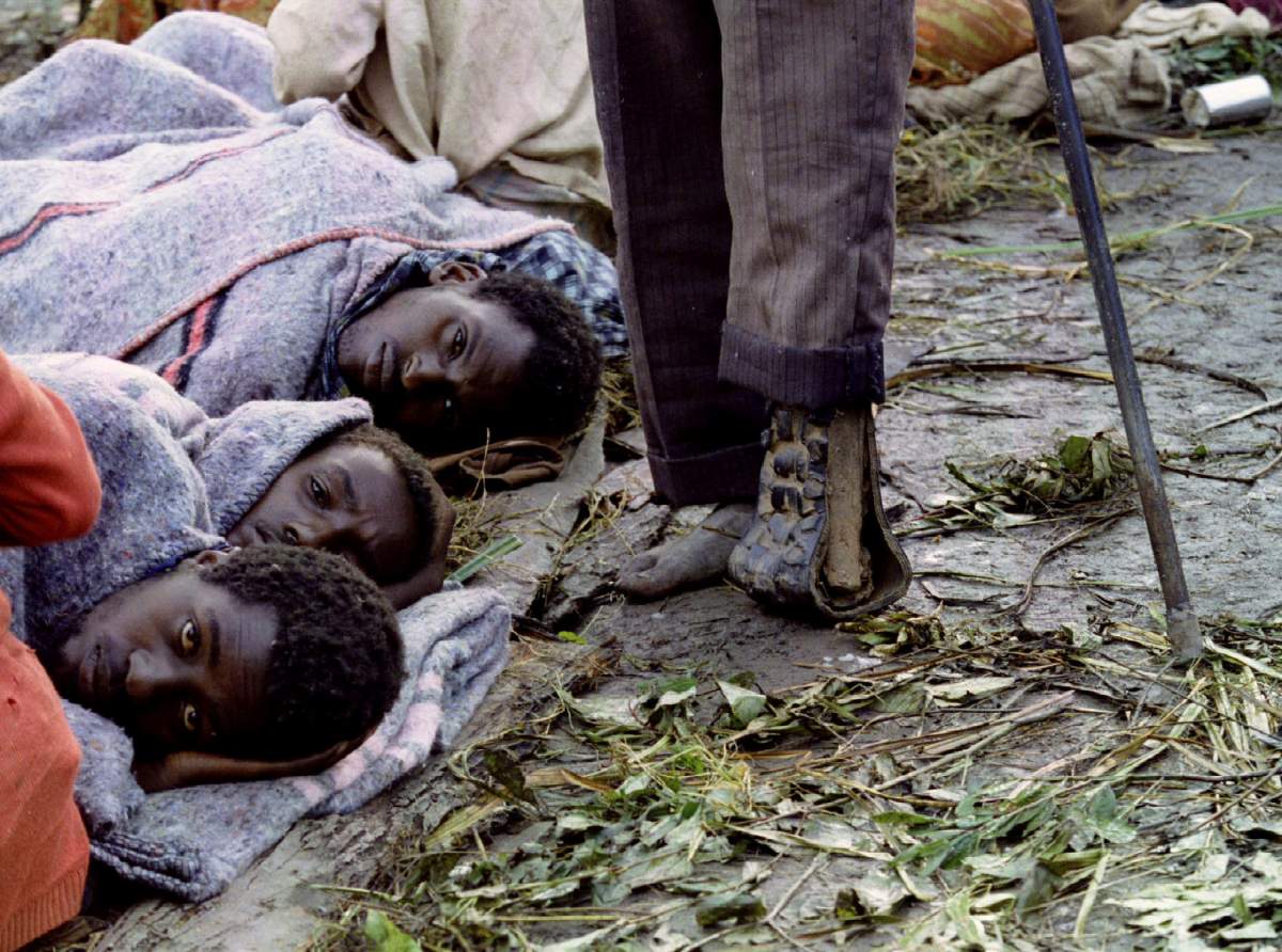 An amputee moves past three Tutsi refugees huddled together to protect themselves against the cold and damp in this camp in southern Rwanda, May 20 1994.