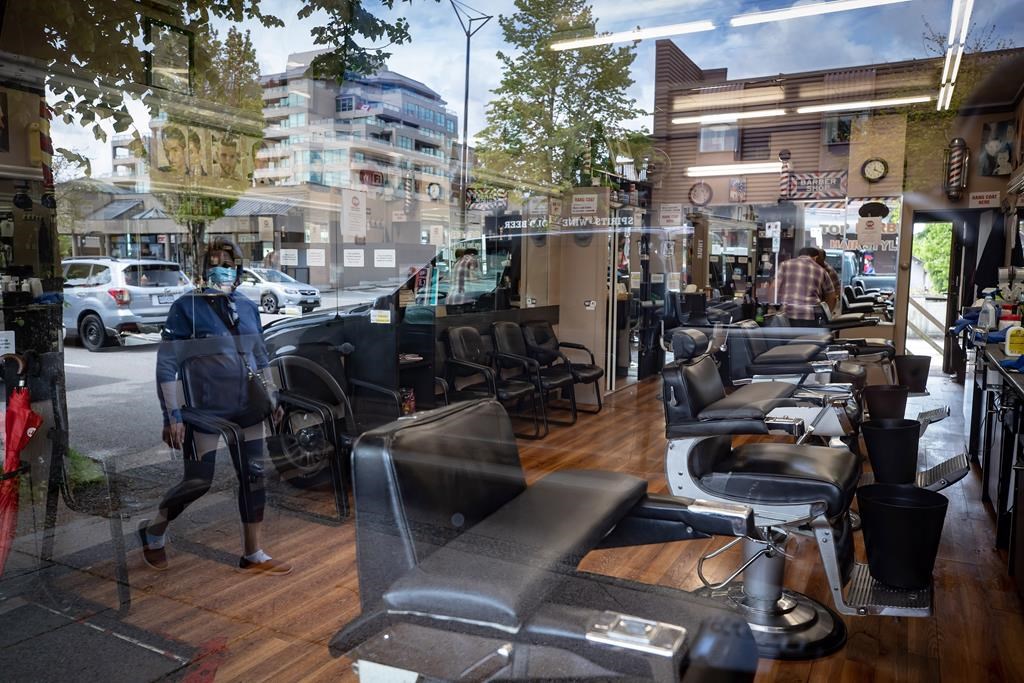 A man cleans inside a closed barbershop as a pedestrian wearing a face mask is reflected in the shop's window, in Burnaby, B.C., on Wednesday, May 6, 2020. Salons and barbershops are starting to reopen in some parts of Canada with more on the way, but the industry says that the measures needed to keep everyone safe while getting a haircut will mean a big financial hit. THE CANADIAN PRESS/Darryl Dyck.