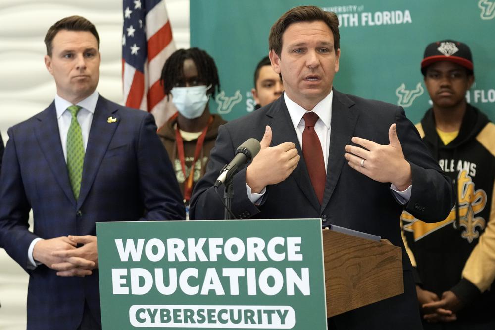 Ron DeSantis stands behind a lectern wearing a suit and tie at a press conference. Students stand behind him.