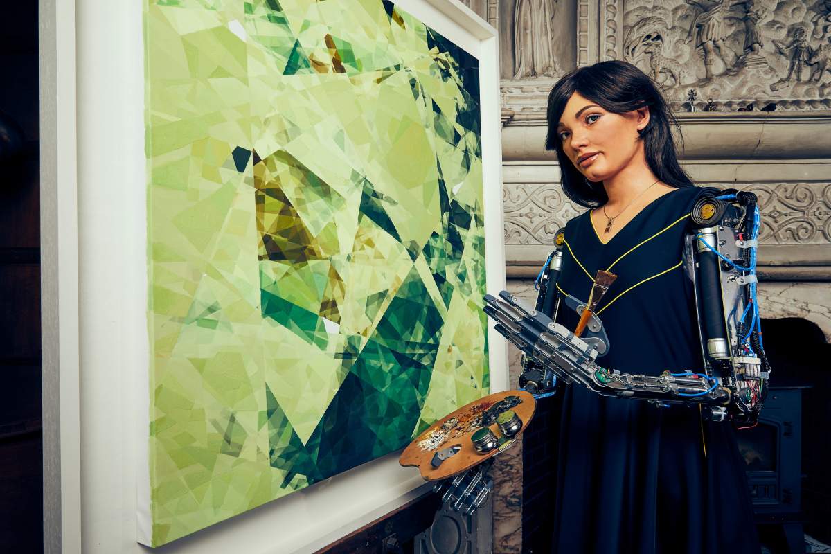 ai-da stand beside an original work of art. She wears a dress and has a very realistic face, with long brown hair, but her robotic arms are fully exposed.
