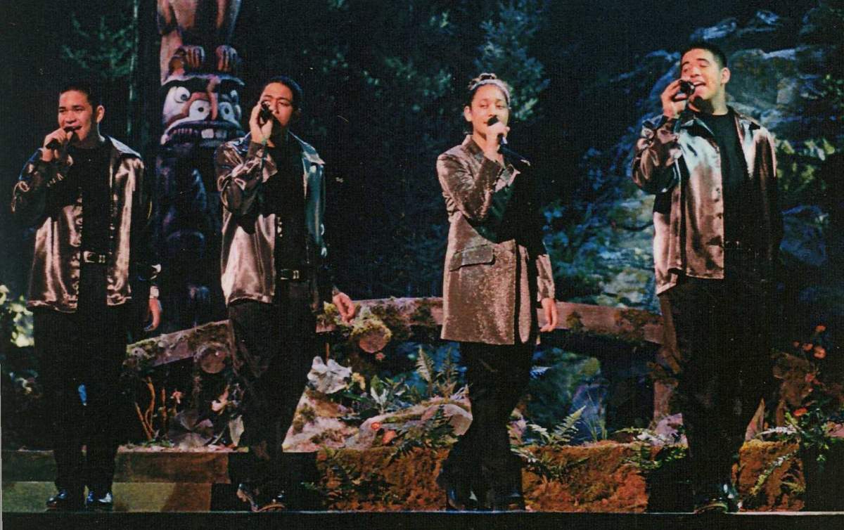 Four members of the pop-group, The 5th Generation, holding wireless microphones while perrming on stage for a live audience at an Indigenous award show in 1999. They are wearing matching outfits of a shiny jacket and black blanks.
