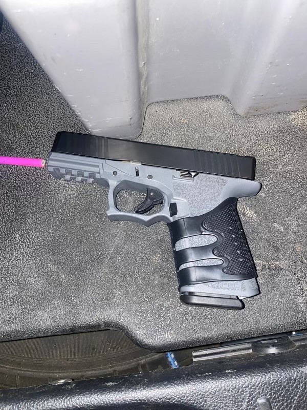 A handgun laid on the floor. Beside the barrel is the tip of a pink coloured pencil.