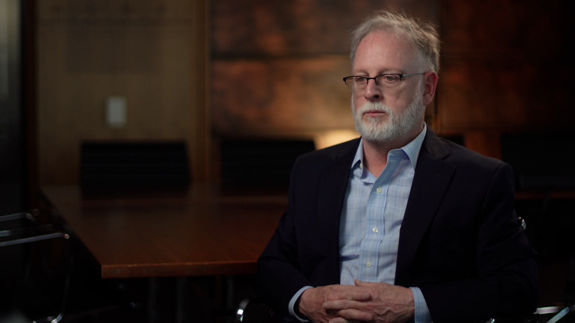 Bob Kirk, a man with a white beard and white hear, is looking to the left of the screen. He is wearing glasses, a blue collared shirt, and a blazer.