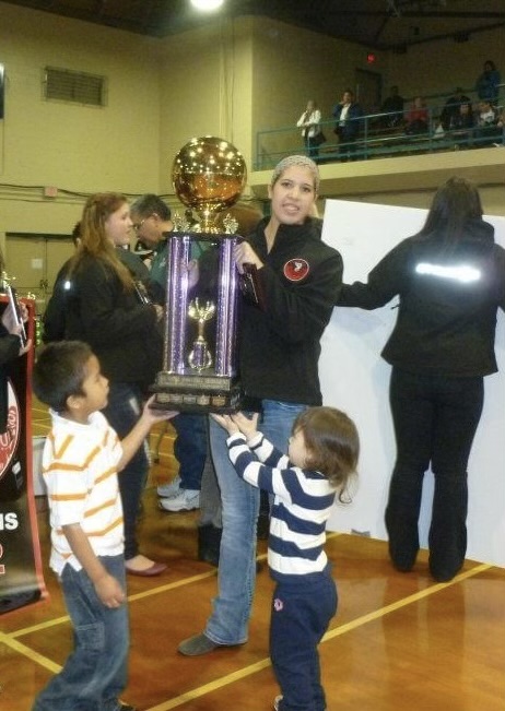 A woman stands holding a large trophy with two children in front of her reaching for the trophy.