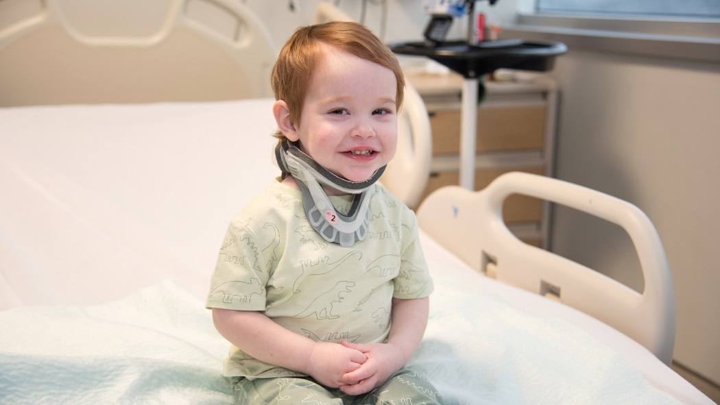A toddler in a neck brace smiles while sitting on a hospital bed.