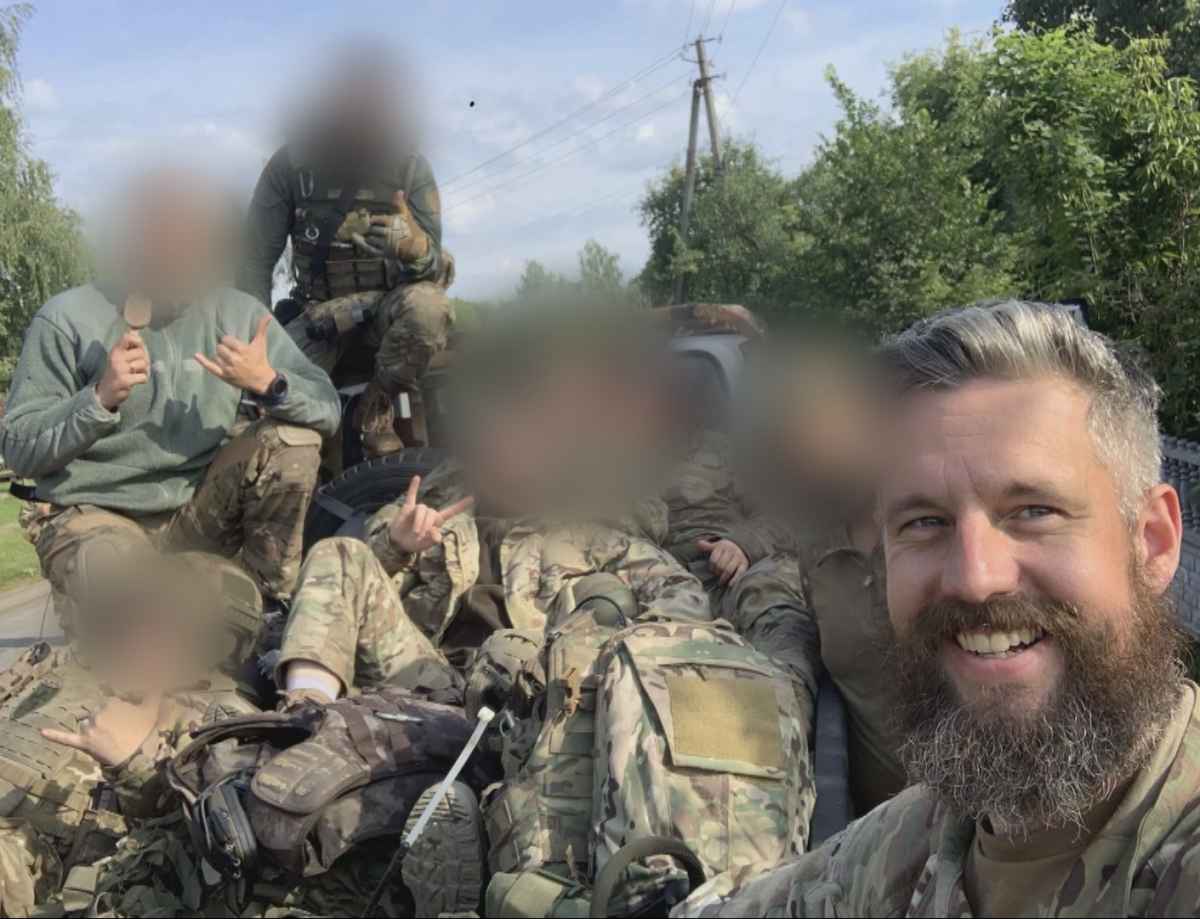 A group of soldiers smile for a selfie while seated in a pickup truck in Ukraine. The faces of all but one - Canadian Dave Smith - are blurred.