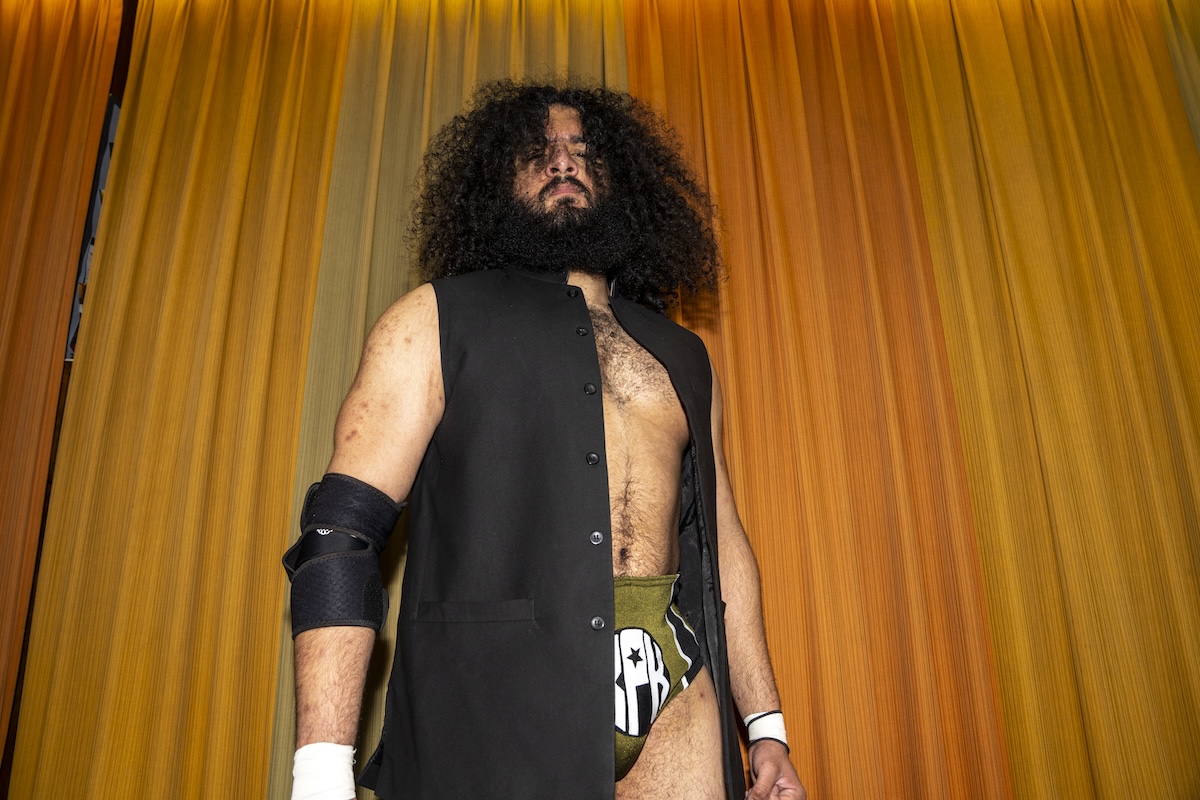Man with black curly hair wearing a black vest and green briefs stands in front of an orange and green curtain backdrop.