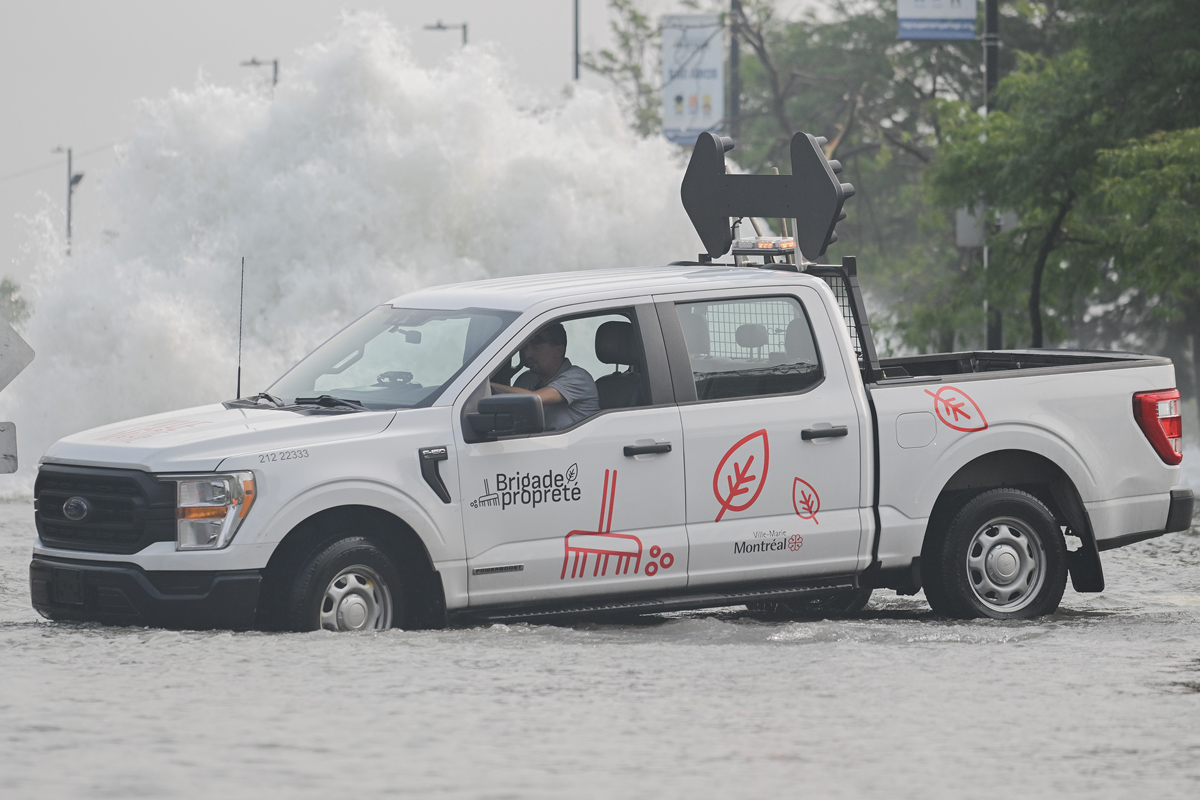 A city vehicle navigates a flooded street next to a geyser caused by a broken watermain in Montreal, Friday, August 16, 2024, causing flooding in several streets of the area.