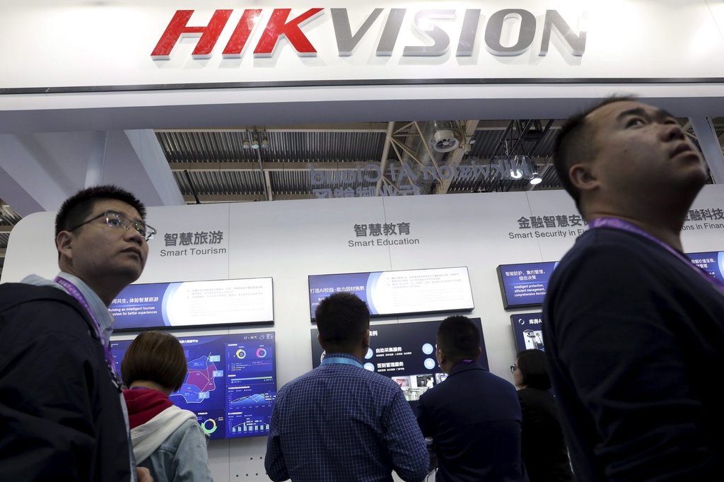 In this Tuesday, Oct. 23, 2018, file photo, visitors pass by a booth for state-owned surveillance equipment manufacturer Hikvision at the Security China 2018 expo in Beijing, China. (AP Photo/Ng Han Guan, File).