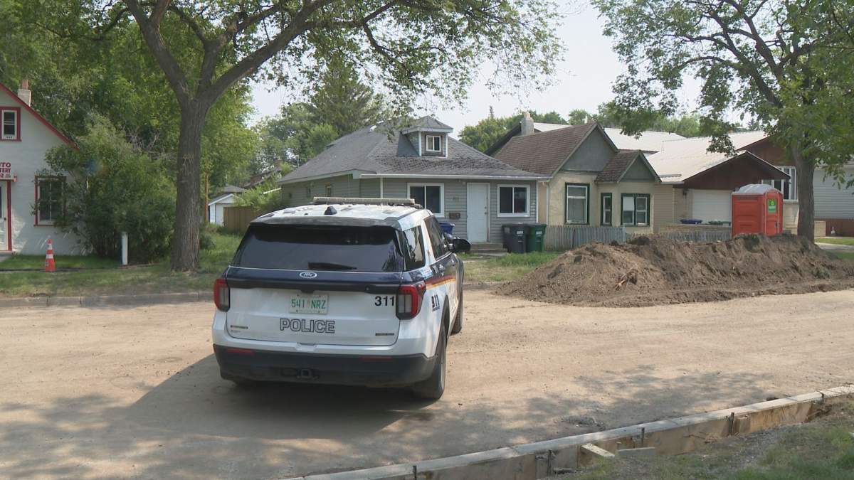 A Saskatoon police cruiser blocks a street next to a dirt pile, with a one-storey home in the background framed by large trees.