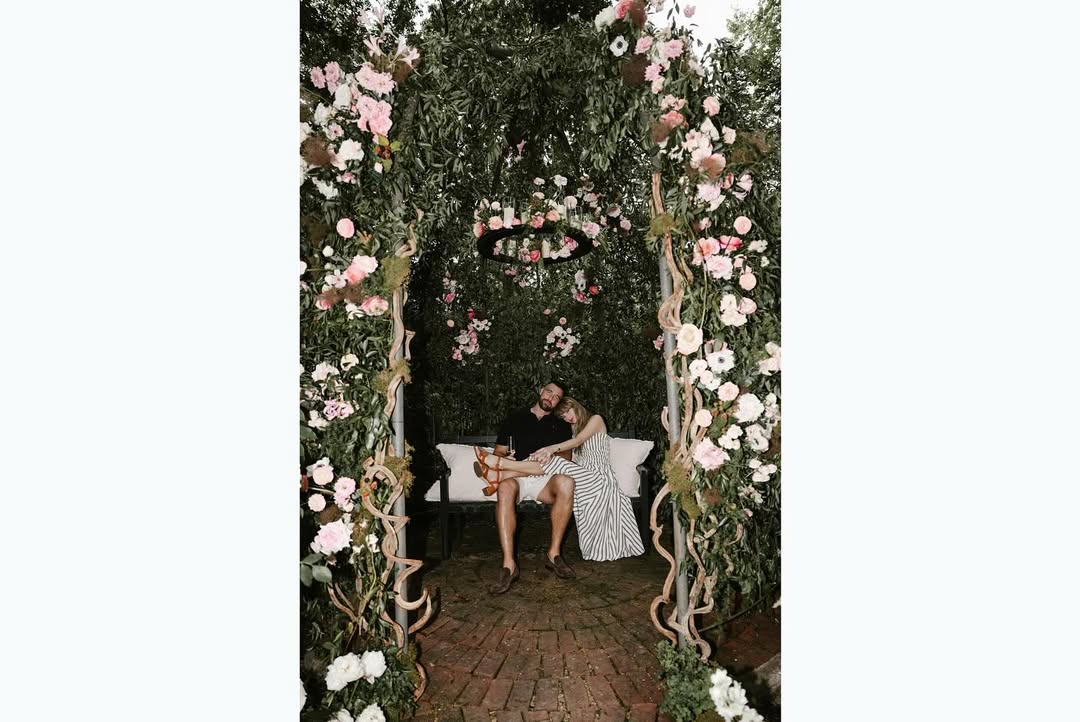 The last picture in the slide show is of the couple sitting on a bench beneath an archway full of flowers on trellises. Swift is resting her head on Kelce’s shoulder, with her legs across his lap. She wears brown sandals and Kelce is dressed in a pair of white shorts.