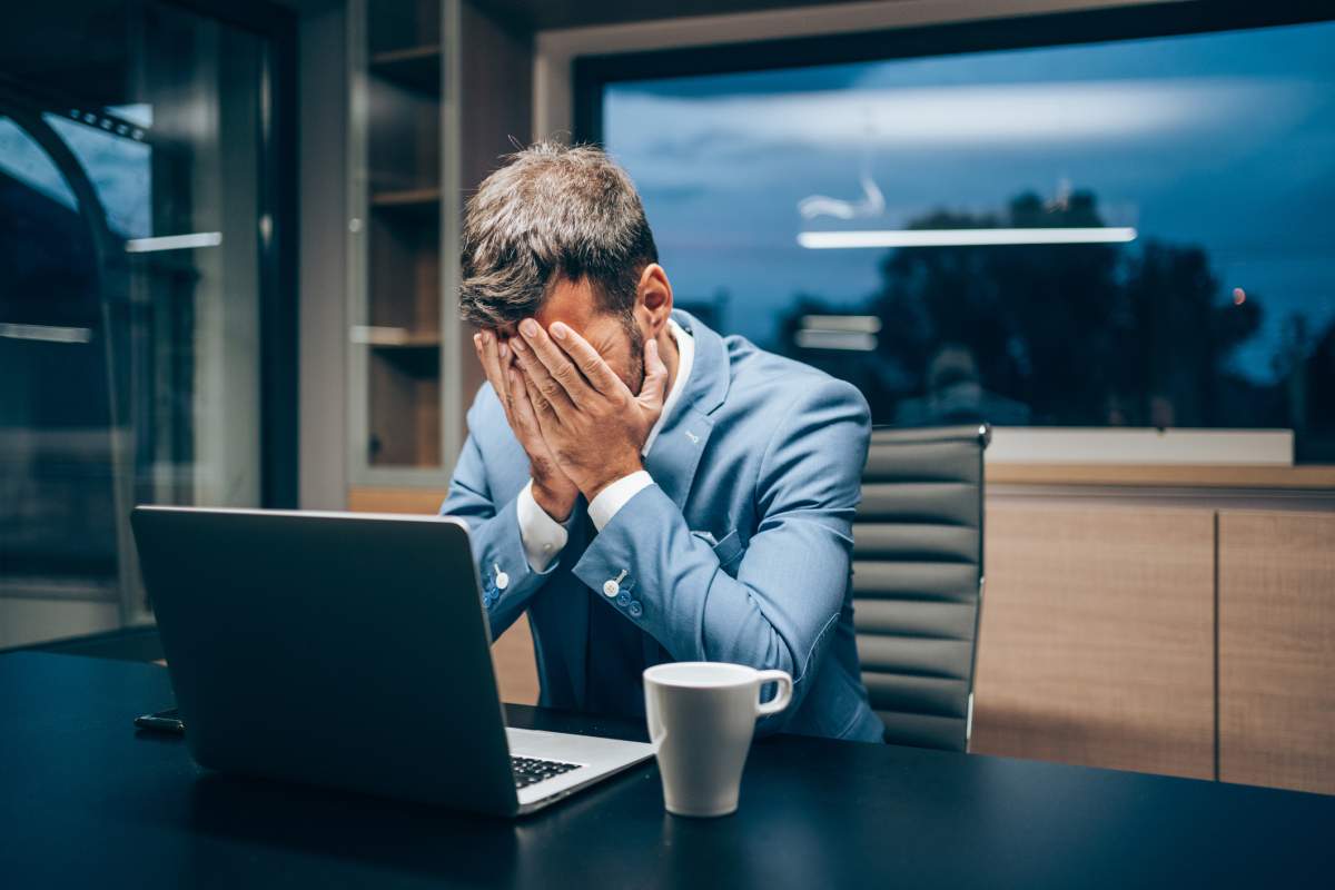A businessman covers his face while sitting in front of a laptop.