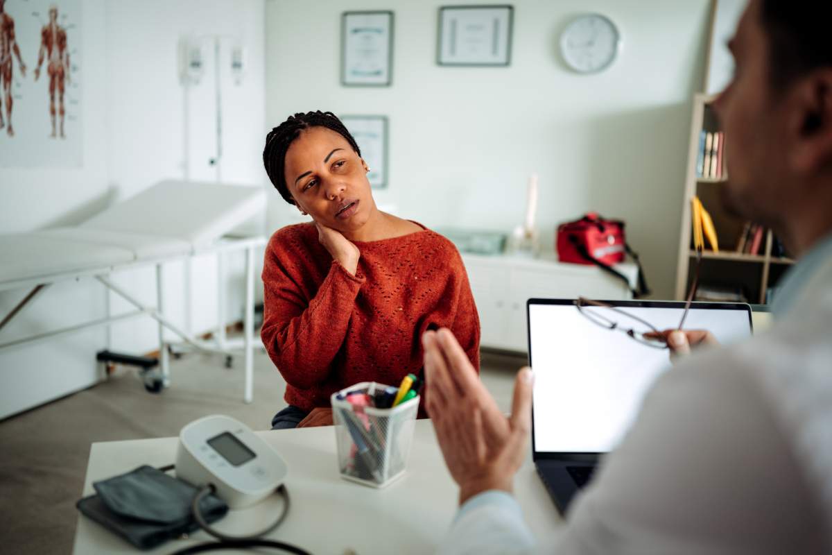 A woman rubs her neck while listening to her doctor during an appointment. Mixetto/Getty Images