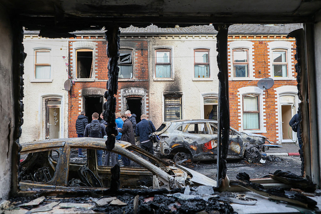 People stand next to burnt-out cars and homes after demonstrations turned violent the night before, in eastern Belfast, Northern Ireland, on June 10, 2026. A Sudanese man was due to appear in court in Northern Ireland on June 10 over a brutal knife attack captured on video that triggered a night of violence by anti-immigration protesters.
